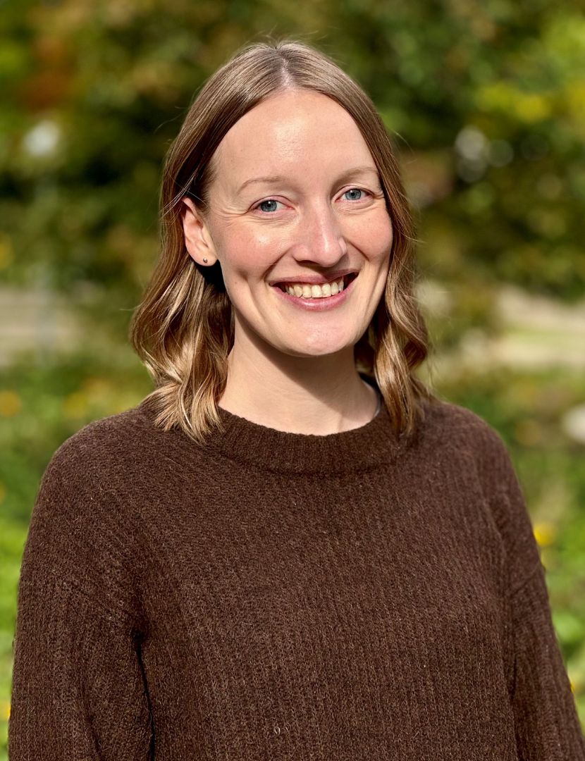 Melody Smale smiling in front of a blurred outdoor background. She has light brown hair that reaches her shoulders and is wearing a brown knit sweater.